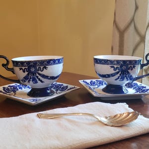 May include: Two white and blue teacups with matching saucers. The teacups have a blue ribbon design and gold trim. The handles are dark blue. A silver spoon rests on a white cloth.