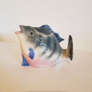 以下が含まれることがあります： 口が開いた注ぎ口を持つ魚の形をした陶器製のピッチャー。胴体は背中側が濃い青とグレーの鱗のグラデーションになっており、腹部にかけて淡いピンク色に変化しています。目立つ背びれと暗い色の尾びれが付いています。