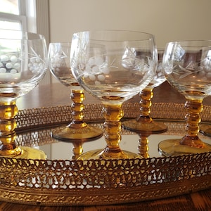 May include: A set of six vintage wine glasses with clear bowls and amber-colored stems. Each glass features an etched grape and vine design. The glasses are arranged on a gold-toned mirrored tray with a decorative edge, reflecting the glasses.