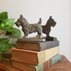 May include: A pair of bronze-toned metal Scottish Terrier dog bookends featuring textured fur details, standing on rectangular bases. The bookends are positioned on top of a stack of three hardcover books with visible titles on the spines.