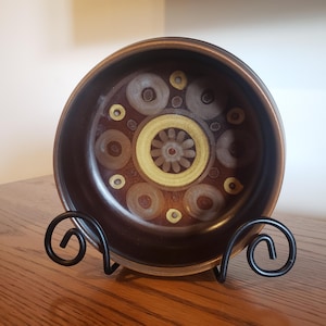 May include: A brown ceramic bowl with a decorative floral and circular pattern in shades of gold and brown. The bowl is displayed on a black metal stand, showcasing its design. The bowl is a decorative piece.