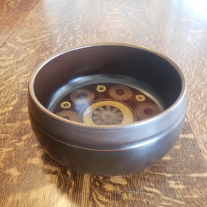 May include: A brown ceramic bowl with a decorative interior. The interior features a circular design with concentric circles in shades of brown, tan, and yellow. The bowl has a smooth, rounded exterior and a slightly flared rim.