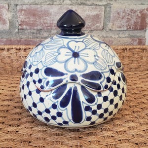 May include: A ceramic jar with a lid, decorated with white and blue floral patterns. The jar has a rounded shape and a dark blue finial. The design includes stylized flowers and dots, creating a decorative piece. The jar is approximately 15 cm in diameter.
