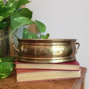 May include: An oval brass bowl with a polished metallic finish and two small side handles. The bowl features a smooth, rounded rim and a slightly tapered base, resting on top of two stacked books with red covers and visible page edges.