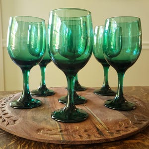 Op de afbeelding: Een set van zes smaragdgroene wijnglazen. De glazen hebben een klassiek ontwerp met een afgeronde kom en een lange steel. Ze zijn gerangschikt op een ronde, houten serveerschaal met gesneden details. De glazen zijn leeg.