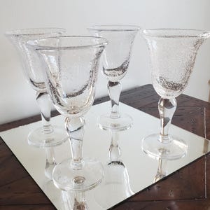 May include: Four clear, stemmed wine glasses with a textured, bubble-like pattern on the bowl. The glasses are displayed on a mirrored surface, reflecting their shape. The stems are slender, and the bases are round.