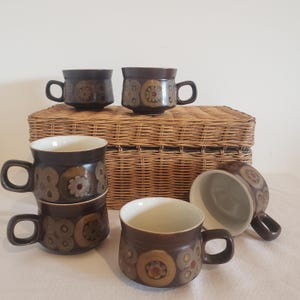 May include: A set of six brown ceramic mugs with floral and geometric patterns. The mugs have a dark brown exterior, white interior, and rounded handles. The mugs are arranged on a white surface with a wicker basket in the background.