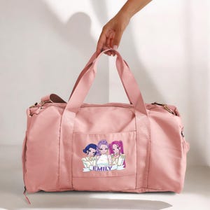 May include: A pink duffel bag with a shoulder strap and handles. The bag features a graphic of three anime-style girls with the name "EMILY" below. The bag is held by a hand.