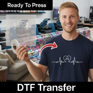 May include: A navy blue t-shirt with a white graphic of a bicycle inside a heart, with a heartbeat line. The image also shows a clear transfer sheet with the same design. The text "Ready To Press" and "DTF Transfer" are visible.