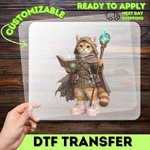 May include: A DTF transfer sheet featuring a ginger cat dressed as a wizard in a hooded cloak, holding an open glowing book and a staff topped with a luminous orb. The cat wears a belt with pouches and light pink boots. Text reads CUSTOMIZABLE.