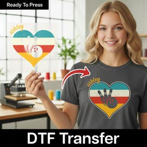 May include: A dark gray t-shirt featuring a heart-shaped graphic with horizontal stripes in teal, white, red, and yellow. The graphic includes a bowling ball and pins with the name ashley written in yellow cursive script above the heart.