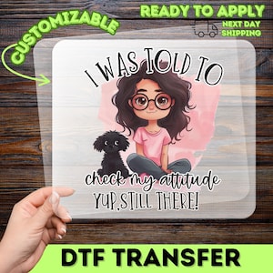 May include: A clear DTF transfer sheet featuring a cartoon illustration of a person with dark wavy hair and glasses sitting next to a small black dog. The text reads: I WAS TOLD TO check my attitude YUP, STILL THERE! The design has a pink watercolor background.