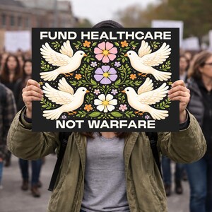 May include: A black sign with the words "FUND HEALTHCARE NOT WARFARE" in white. The sign features four white doves with floral and leaf designs in pink, purple, and yellow. The sign is held up in a crowd of people.