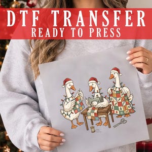 May include: A DTF transfer sheet featuring a graphic of three white geese wearing Santa hats and Christmas lights. One goose sews a quilt, another uses a sewing machine, and the third holds a finished quilt. The text reads DTF TRANSFER READY TO PRESS.