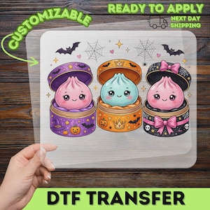 May include: A DTF transfer sheet featuring three cartoon dumplings in round containers. The left is purple with pumpkins and bats, the center is orange with crowns, and the right is black with pink bows and skulls. Bats and spiderwebs decorate the top.
