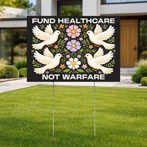 May include: A black yard sign with the text "FUND HEALTHCARE NOT WARFARE" in white. The sign features four white doves and floral designs in pink, purple, and orange. The sign is displayed on a metal stake in a grassy yard.