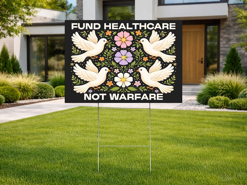 May include: A black yard sign with the text "FUND HEALTHCARE NOT WARFARE" in white. The sign features four white doves and floral designs in pink, purple, and orange. The sign is displayed on a metal stake in a grassy yard.