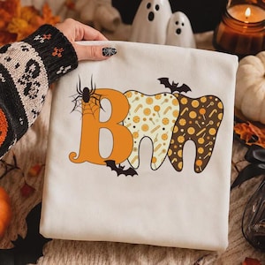 May include: A white sweatshirt featuring a graphic of the letters B, O, and O shaped like a tooth. The B is orange with a black spider and web. The O letters are filled with a pattern of orange and yellow candies, accented by black bats.
