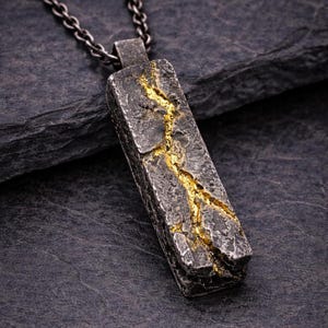 Op de afbeelding: Een rechthoekige hanger met een getextureerd, donkergrijs oppervlak en een goudkleurige scheur die door het midden loopt. De hanger hangt aan een zilverkleurige ketting. Het sieraad heeft een rustieke, industriële uitstraling.