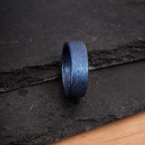 May include: A blue ring with a speckled, glittery finish. The ring is set against a dark backdrop, emphasising its colour and texture. The ring is a piece of jewellery.