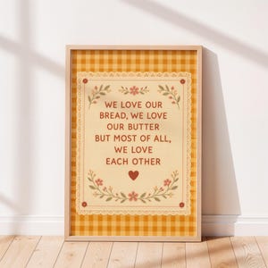 May include: A framed print with a yellow and white gingham background. The print features a cream-colored rectangle with the text "WE LOVE OUR BREAD, WE LOVE OUR BUTTER BUT MOST OF ALL, WE LOVE EACH OTHER" in brown, with floral accents.
