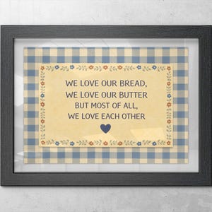 May include: Framed print with a black frame. The print features a cream-colored background with text that reads "WE LOVE OUR BREAD, WE LOVE OUR BUTTER BUT MOST OF ALL, WE LOVE EACH OTHER." The text is surrounded by a blue and white gingham pattern.