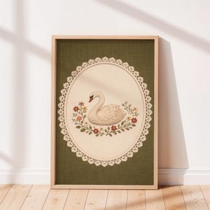 May include: Framed artwork featuring a white swan with a red beak, surrounded by colorful flowers and green foliage. The swan is set within an oval frame with a lace-like border, against a green background. The artwork is encased in a light brown wooden frame.
