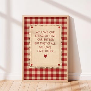 May include: Framed print with a red and cream gingham pattern background. The print features a cream-colored center with the text "WE LOVE OUR BREAD, WE LOVE OUR BUTTER BUT MOST OF ALL, WE LOVE EACH OTHER" and a red heart.