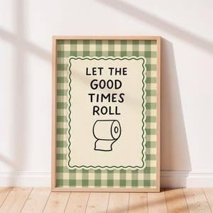 Puede incluir: Un cuadro enmarcado con el texto "LET THE GOOD TIMES ROLL" y una ilustración de un rollo de papel higiénico. El fondo presenta un patrón de cuadros verdes y blancos, y el marco es de madera clara.