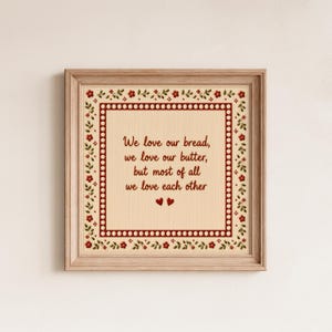 Könnte beinhalten: Ein gerahmtes Kunstwerk mit einem hellen Holzrahmen. Das Kunstwerk zeigt einen cremefarbenen Hintergrund mit dem Text "We love our bread, we love our butter, but most of all we love each other." Roter Blumen- und Blätterrand.