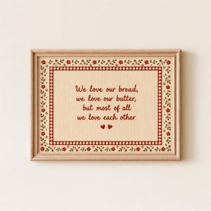 May include: Framed artwork with a light beige background and a decorative red border. The text reads "We love our bread, we love our butter, but most of all we love each other" in red script. Two red hearts are at the bottom.