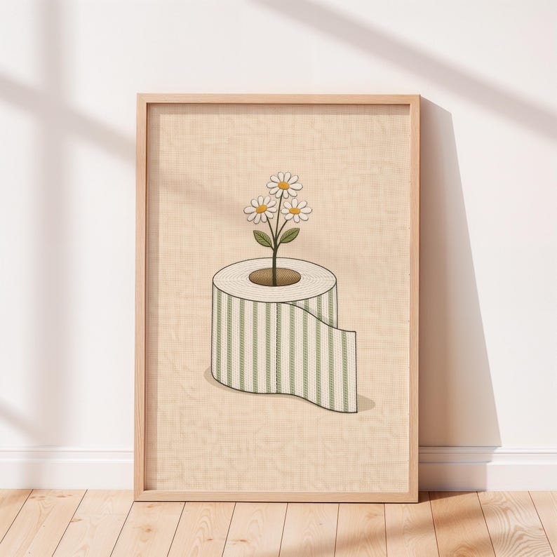 May include: A framed art print featuring a roll of toilet paper with green and white stripes, with a daisy plant growing from the center. The artwork has a light beige background and is framed in a light wood color.