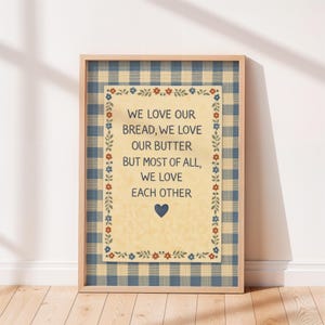 May include: A framed print with a light wood frame. The print has a blue and white gingham border and a cream-colored central panel. The text reads "WE LOVE OUR BREAD, WE LOVE OUR BUTTER BUT MOST OF ALL, WE LOVE EACH OTHER" with a blue heart.