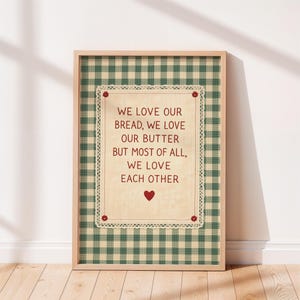 May include: A framed print with a light wood frame. The print features a cream-colored rectangle with the text "WE LOVE OUR BREAD, WE LOVE OUR BUTTER BUT MOST OF ALL, WE LOVE EACH OTHER" in red. The background is green and white checkered.
