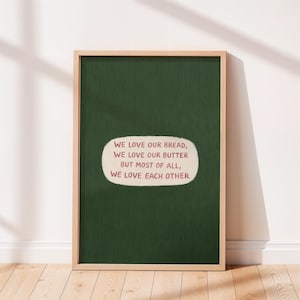 Pode incluir: Uma impressão artística emoldurada com um fundo verde escuro e uma moldura de madeira clara. A impressão apresenta o texto "WE LOVE OUR BREAD, WE LOVE OUR BUTTER BUT MOST OF ALL, WE LOVE EACH OTHER" em um oval branco com texto vermelho.