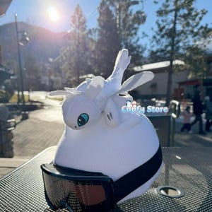 Peut inclure: Un bonnet blanc en tricot avec une bande noire et des lunettes de protection attachées. Un dragon en peluche blanc avec des yeux bleus et des ailes blanches est posé dessus. Le texte "Capfy Store" est visible.