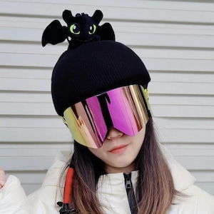 May include: A black knit beanie featuring a plush black dragon character with green eyes and wings attached to the top. The beanie is paired with large, frameless ski goggles that have a reflective, iridescent pink and gold lens finish.