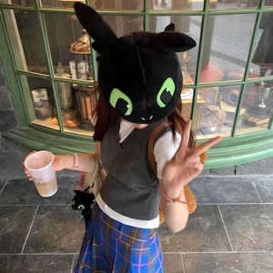 Peut inclure: Un chapeau de dragon noir avec des yeux et des oreilles verts. La personne porte un gilet gris, une chemise blanche et une jupe à carreaux bleue. La personne tient une tasse avec le mot "Potter" dessus.