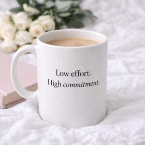 Puede incluir: Taza de cerámica blanca llena de café, con el texto "Low effort. High commitment." Un libro rosa y flores blancas están en el fondo. La taza tiene un asa curva y está sobre una superficie blanca.