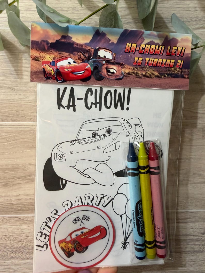 May include: A clear plastic bag containing a colouring book and crayons. The colouring book features a black and white outline of a race car with the text "KA-CHOW!" and "LET'S PARTY". The bag also includes blue, yellow, and pink crayons.