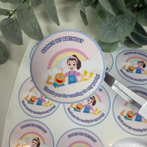 May include: A sheet of circular stickers featuring a cartoon illustration of a child and a yellow monster character under a rainbow. The text reads Name's 1st Birthday and Thank you for coming to my party in blue lettering on a light pink background.
