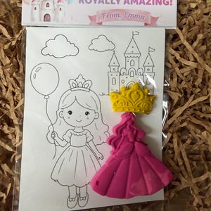 Peut inclure: Un sachet plastique transparent contenant une feuille de coloriage avec un dessin au trait d'une princesse, d'un château et d'un ballon. Un crayon jaune en forme de couronne et un crayon rose en forme de princesse sont fixés devant. L'en-tête indique YOU'RE A ROYALLY AMAZING! From: Emma.