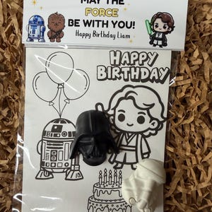Peut inclure: Un sachet plastique transparent contenant une carte blanche avec des dessins au trait noir représentant un robot, un personnage, des ballons et un gâteau avec le texte "HAPPY BIRTHDAY". Un casque de Dark Vador noir et une figurine de stormtrooper blanche sont fixés sur la carte. L'en-tête indique "MAY THE FORCE BE WITH YOU! Happy Birthday Liam" avec des illustrations.