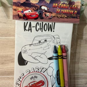May include: A clear plastic bag containing a colouring book and crayons. The colouring book features a black and white illustration of a race car with the text "KA-CHOW!" and "LET'S PARTY". The bag also includes blue, yellow, and red crayons. The top of the bag has a banner with the text "HA-CHOW! LEVI IS TURNING 21!"