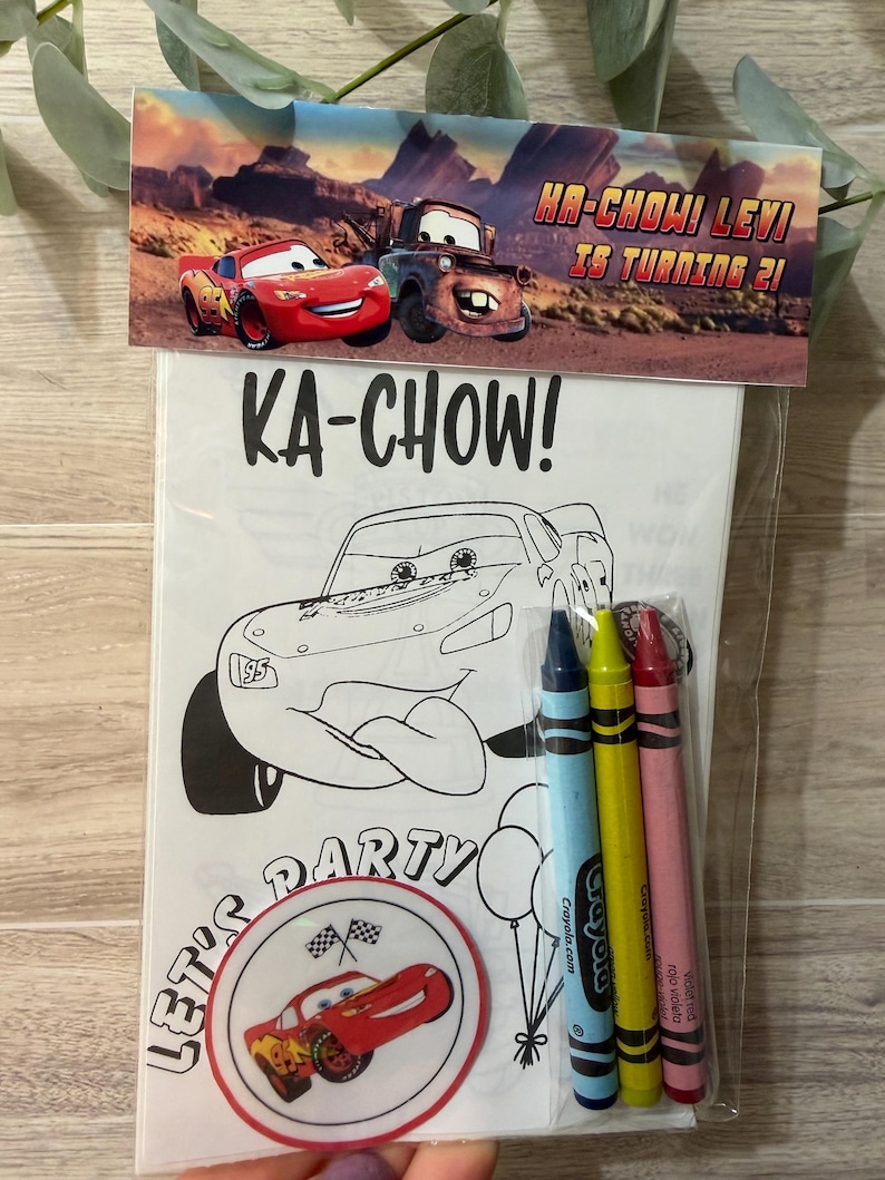 May include: A clear plastic bag containing a colouring book and crayons. The colouring book features a black and white illustration of a race car with the text "KA-CHOW!" and "LET'S PARTY". The bag also includes blue, yellow, and red crayons. The top of the bag has a banner with the text "HA-CHOW! LEVI IS TURNING 21!"