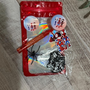 May include: A red cellophane bag filled with Spider-Man themed accessories. Includes stickers, a glitter pen, a plastic spider, and a small black item. Two round stickers say "Thank You".