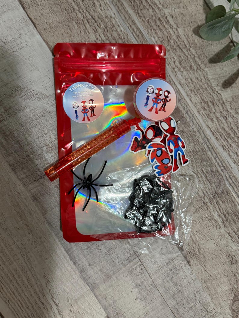 May include: A red cellophane bag filled with Spider-Man themed accessories. Includes stickers, a glitter pen, a plastic spider, and a small black item. Two round stickers say "Thank You".