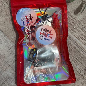 May include: A red cellophane bag filled with Spider-Man themed party favors. The bag contains stickers, a pen, a plastic spider, and two circular stickers that read "Thank you for celebrating my birthday with me!"