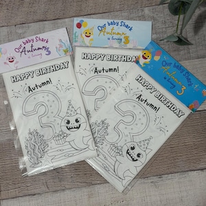 Peut inclure: Trois pages à colorier sur le thème de Baby Shark dans des pochettes en plastique. Chaque page indique "Joyeux Anniversaire Autumn !" et présente un grand chiffre 3 avec un requin portant un chapeau de fête. La bannière supérieure indique "Our baby Shark Autumn is turning 3".