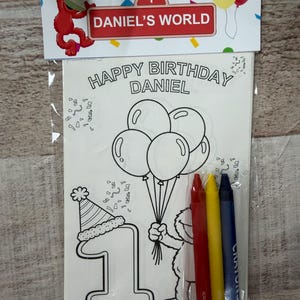May include: A clear plastic package containing a coloring sheet and crayons. The sheet says "Happy Birthday Daniel" and features a cartoon character holding balloons. Includes red, yellow, and blue crayons. The package is labeled "Daniel's World".